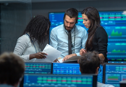 a small group of people talking on a trading floor with computer screens and large screens showing numbers surrounding them